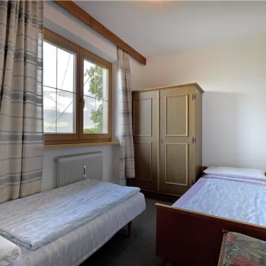 A simple bedroom with two single beds and a wardrobe. Large windows let in plenty of natural light.