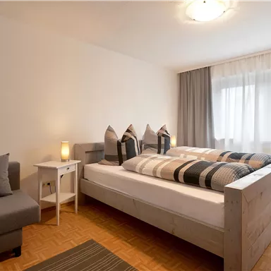 A modern bedroom with a large bed and cozy pillows. There is a seating area and plenty of daylight through the windows.