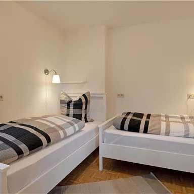 A cozy bedroom with two single beds and modern reading lamps. The walls are neutral, and there are decorative cushions with a checkered pattern.