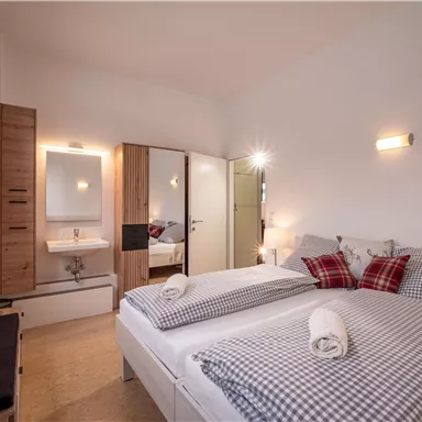 A cozy bedroom with two beds and checkered bedspreads. There is a mirror, a small washing area, and warm lighting.