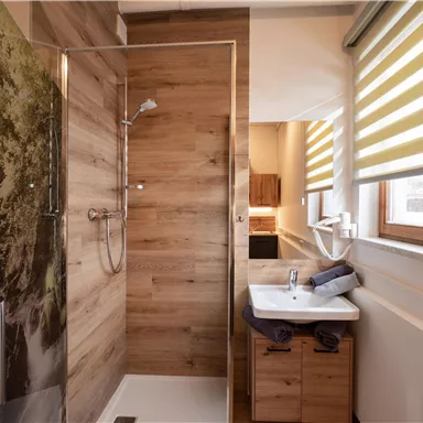 A modern bathroom with a shower and wooden paneling. There is a sink and large windows with blinds that provide plenty of light.