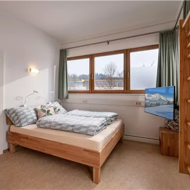 A modern bedroom with a wooden double bed and a television. Large windows offer a view of the landscape and let in plenty of light.