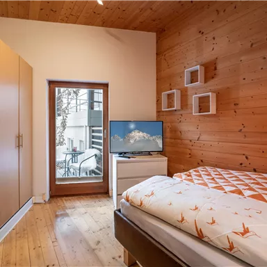 A cozy bedroom with wood paneling, a television, and a window overlooking a balcony. The decor is modern and inviting.