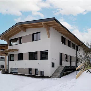 A modern residential building in a snow-covered landscape. The facade is white with large windows and a wooden overhang.