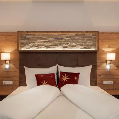 A modern bedroom with a wooden nightstand and a stone wall behind the bed. The beds are decorated with white blankets and red pillows.