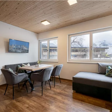 A modern living room with a cozy sofa and a dining area. Large windows provide plenty of daylight.