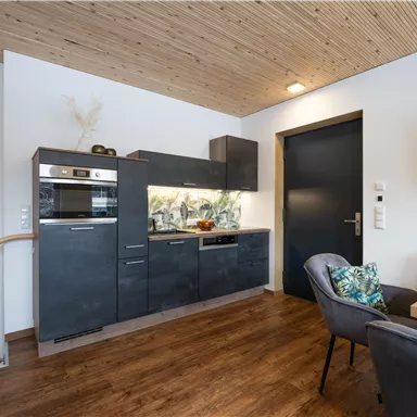 A modern kitchen with black furniture and a wooden ceiling. The space is bright and friendly, with a comfortable dining table.