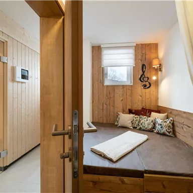 A cozy wellness area with a wooden sofa and a sauna in the background. The walls are made of wood, creating a relaxing atmosphere.