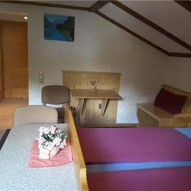 A cozy bedroom with a double bed and wooden decor. There is a table, a chair, and a small flower arrangement on the bedspread.