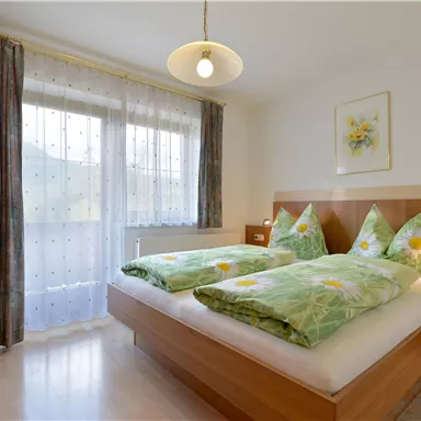 A bright bedroom with two separate beds and colorful bedspreads. Large windows provide plenty of daylight.