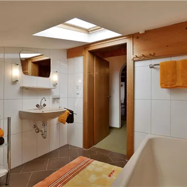 A modern bathroom with a bathtub and a large window. The walls are tiled in white, and orange towels and decorations add accents.