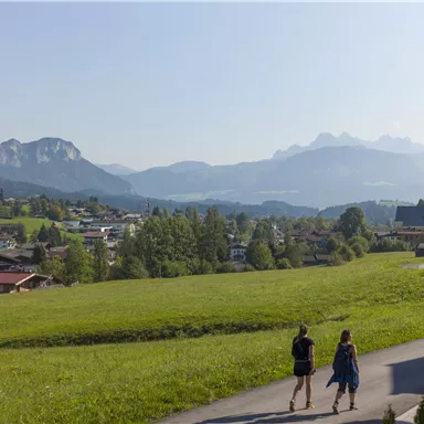 An idyllic landscape with gentle hills and a view of the mountains. Two people are walking near a small village.