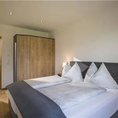 A cozy bedroom with a large bed and elegant bedding. The room is bright and furnished in a minimalist style, with a wooden wardrobe and warm lighting.