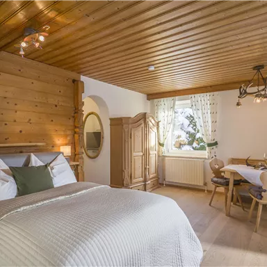A cozy room with a double bed and wooden details. The room has a dining table next to a window overlooking nature.