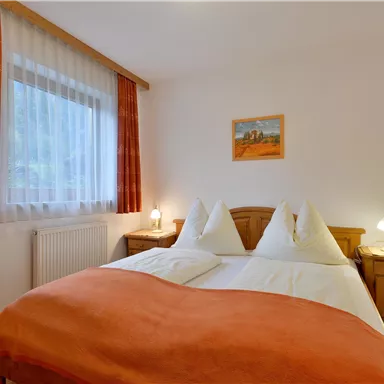 A cozy bedroom with a double bed and an orange bedspread. The walls are light, and natural light comes through the window.
