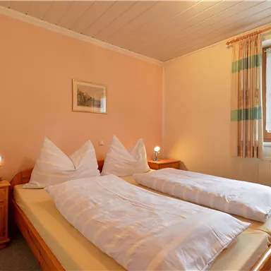 A cozy bedroom with two single beds and bright walls. There are wooden furniture and a window with curtains that lets in natural light.