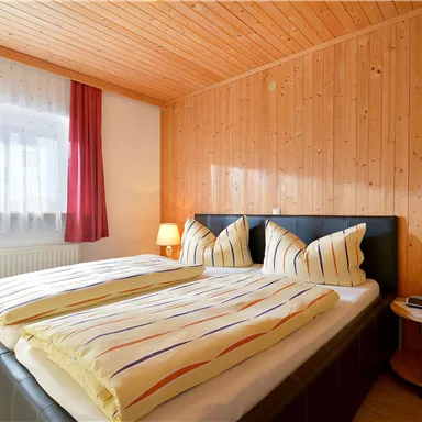 Ein gemütliches Schlafzimmer mit Holzvertäfelung und einem großen Doppelbett. Rote Vorhänge und ein Nachttisch ergänzen die ruhige Atmosphäre.