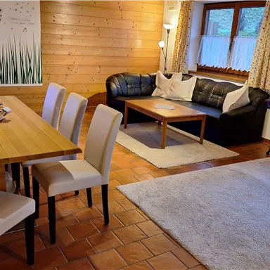 Ein gemütlicher Wohnraum mit einem Esstisch und Stühlen. Im Hintergrund sind ein Sofa und Dekorationen zu sehen.