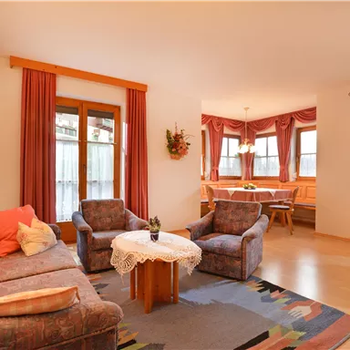 A cozy living room with a sofa and two armchairs. In the background, you can see a dining area and large windows with curtains.