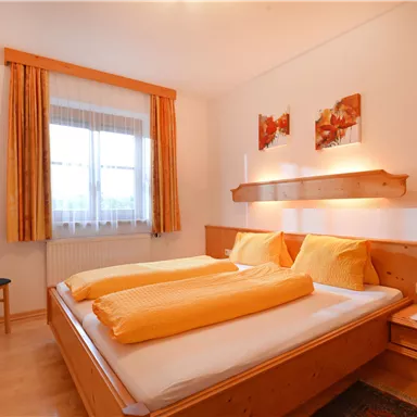 A cozy bedroom with a double bed and two pillows. The room has a window, curtains in warm colors, and a chair in the corner.