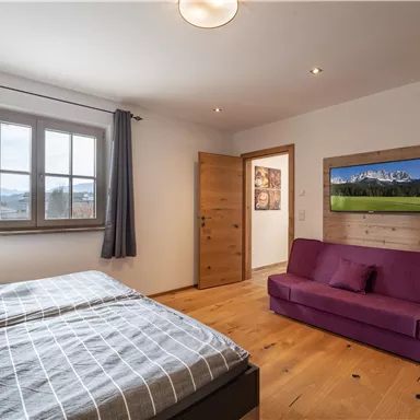 A cozy bedroom with a double bed and a purple sofa. There is a television on the wall and the windows provide plenty of daylight.