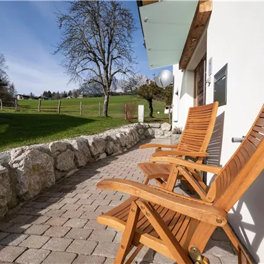 A cozy terrace with two wooden chairs and a view of a green meadow. The sky is clear and the surroundings are peaceful.
