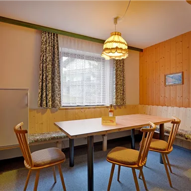 A cozy dining area with wooden walls and a table surrounded by chairs. The windows are decorated with curtains and a lamp hangs from the ceiling.