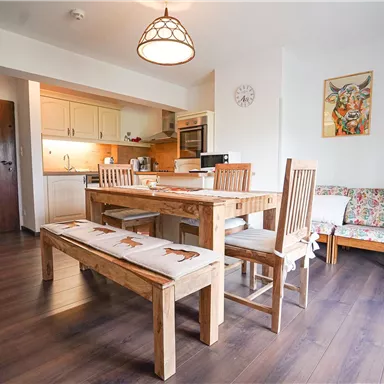 A modern kitchen with a wooden table and chairs. The space is bright and welcoming, with a carpet and artworks on the walls.