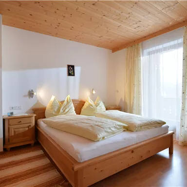 A cozy bedroom with wooden decor. There is a large bed, two pillows, and a chair next to a window with curtains.