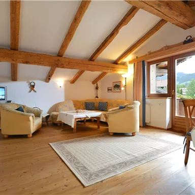 A cozy living room with wooden ceilings and large windows. The room is bright and inviting, with a sofa and a table.