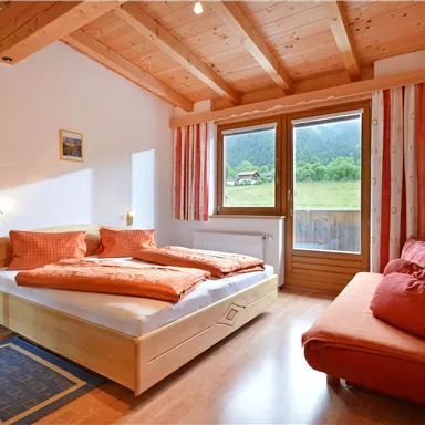 A cozy bedroom with a wooden ceiling and a large window. The room features a double bed with orange pillows and a seating area.