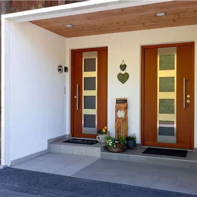 The entrance to a modern house with two wooden doors. Next to the doors stands a decorated plant on the porch.