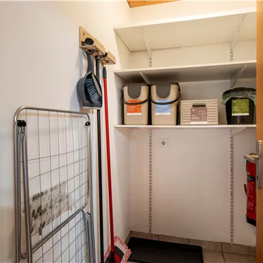A small storage room with shelves and various storage boxes. Cleaning supplies and a coat rack are hanging on the wall.