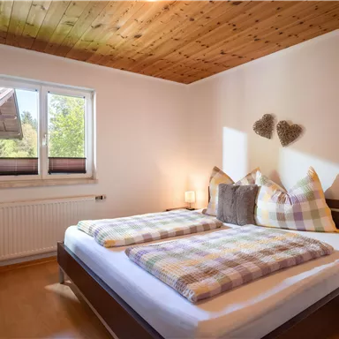 A bright bedroom with a double bed and checkered pillows. Next to the bed is a chair and a window lets in natural light.