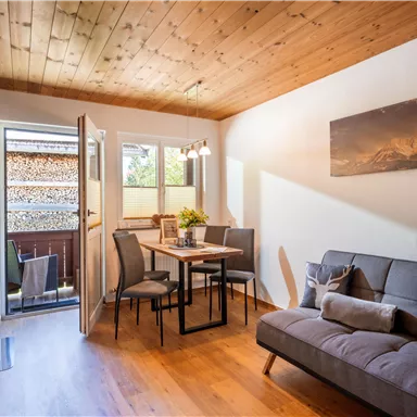 A bright and inviting living kitchen with a dining table and a cozy sofa. Large windows let in plenty of daylight and provide a view of the outdoors.