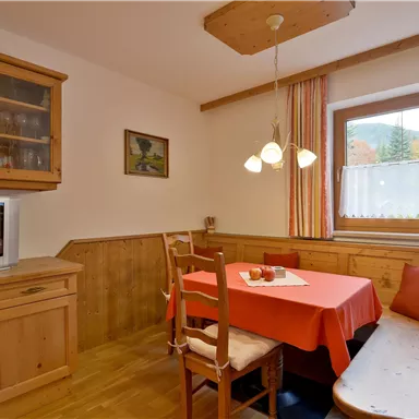 A cozy dining area with a wooden table and chairs. The walls are bright and there are large windows that let in daylight.