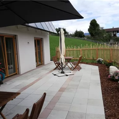 Eine Terrasse mit einem Tisch und Stühlen, umgeben von Blumen und einem Holzzaun. Im Hintergrund sind grüne Wiesen und ein Teil des Himmels sichtbar.