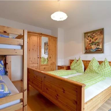 A cozy bedroom with a double bed and a bunk bed. The walls are made of light wood, and there are colorful cushions and a decorative mural.