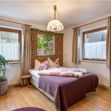 Ein gemütliches Schlafzimmer mit einem Doppelbett, weichen Kissen und natürlichen Holzelementen. Die Wände sind mit einer stilvollen Tapete dekoriert, und große Fenster sorgen für viel Tageslicht.