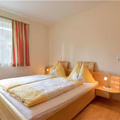 Ein helles Schlafzimmer mit einem Doppelbett und gelben Bettdecken. Große Fenster sorgen für viel Tageslicht und eine einladende Atmosphäre.