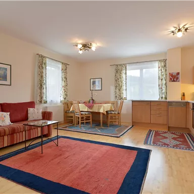 Ein geräumiges Wohnzimmer mit einem roten Sofa und einem Esstisch für vier Personen. Die Küche ist offen und gut ausgestattet, mit großen Fenstern, die viel Licht hineinlassen.