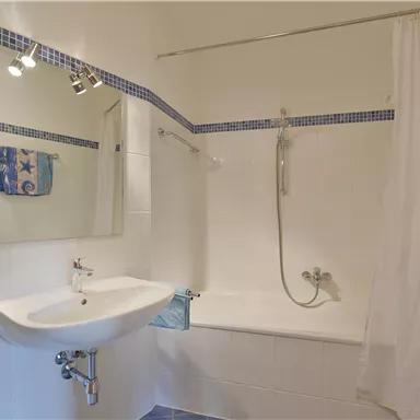 Ein helles Badezimmer mit einer Badewanne und einer Dusche. Am Waschbecken sind Handtücher und ein Spiegel sichtbar.