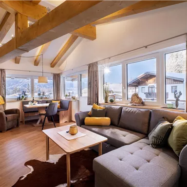 A cozy living room with large windows and a view of the mountains. The wooden ceiling and stylish furnishings create a warm atmosphere.