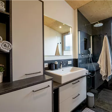 Ein modernes Badezimmer mit dunklen Fliesen und einem hellen Waschbecken. Die Regale sind mit frischen Handtüchern und einer Pflanze dekoriert.