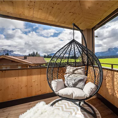 Ein gemütlicher Schaukelstuhl auf einer Terrasse mit Blick auf grüne Wiesen und Berge. Der Himmel ist teilweise bewölkt, was eine entspannende Atmosphäre schafft.