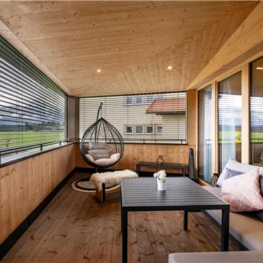 Eine gemütliche Veranda mit Holzverkleidung und großen Fenstern. Ein hängender Stuhl, ein Tisch und bequeme Kissen schaffen eine entspannende Atmosphäre.