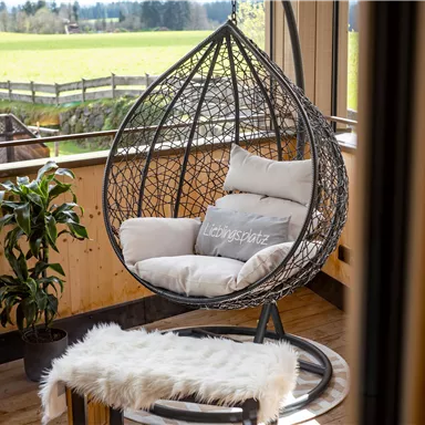 Ein gemütlicher Hängesessel aus Rattan steht in einem Raum mit großem Fenster und Blick auf die Wiesen. Daneben befindet sich eine grüne Pflanze und ein kleiner Tisch mit einem weichen Teppich.