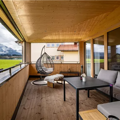 Ein gemütlicher Balkon mit Holzverkleidung, einem Schwingstuhl und Blick auf grüne Wiesen und Berge. Die Sitzgelegenheit lädt zum Entspannen ein.