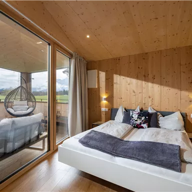 Ein gemütliches Schlafzimmer mit Holzwänden und einem großen Bett. Durch die Fenster hat man Zugang zu einem Balkon mit Ausblick auf die Landschaft.