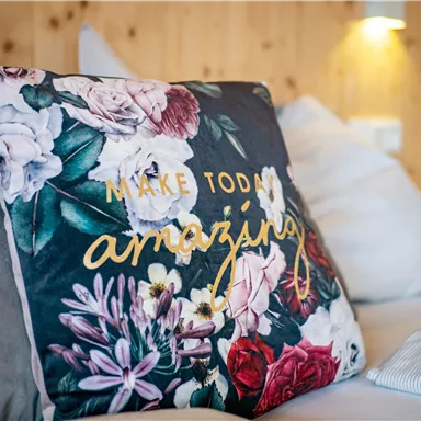 Ein dekoriertes Kissen mit Blumenmuster und der Aufschrift "Make Today Amazing". Es liegt auf einem Sofa mit heller, gemütlicher Umgebung.